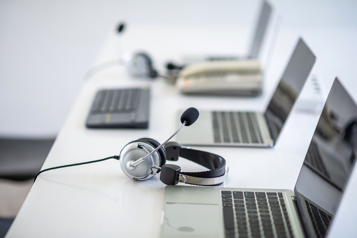 Silver call center headset resting on a white desk beside a row of open laptops and a desk phone in an empty office
