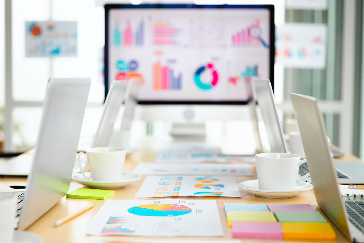 Conference table with laptops, printed charts and sticky notes in front of a monitor showing business analytics