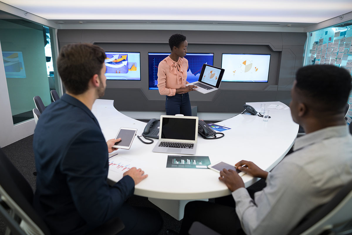 Executive presenting charts on a laptop to two colleagues at a curved white conference table in a high-tech meeting room