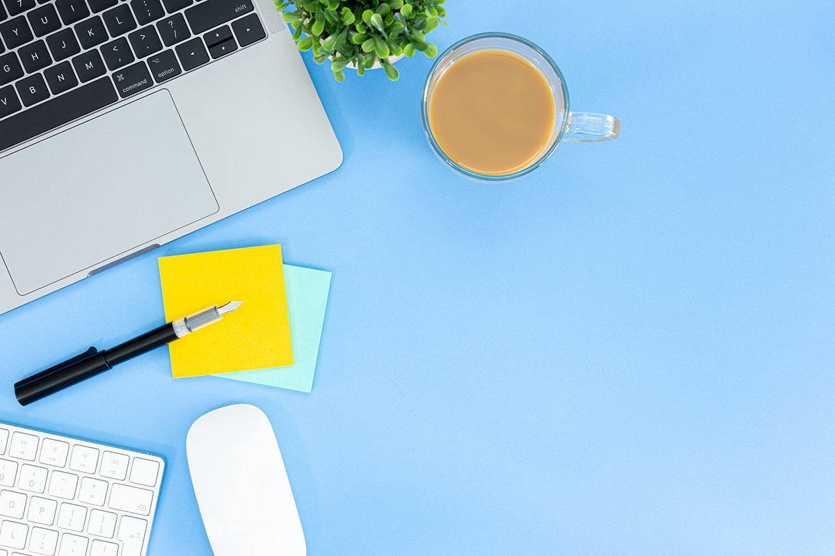 Overhead view of a laptop, keyboard, mouse, fountain pen, yellow sticky notes and a cup of coffee on a light blue desk