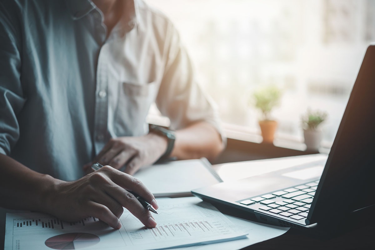 Hand holding a pen over printed financial charts beside an open laptop on an office desk