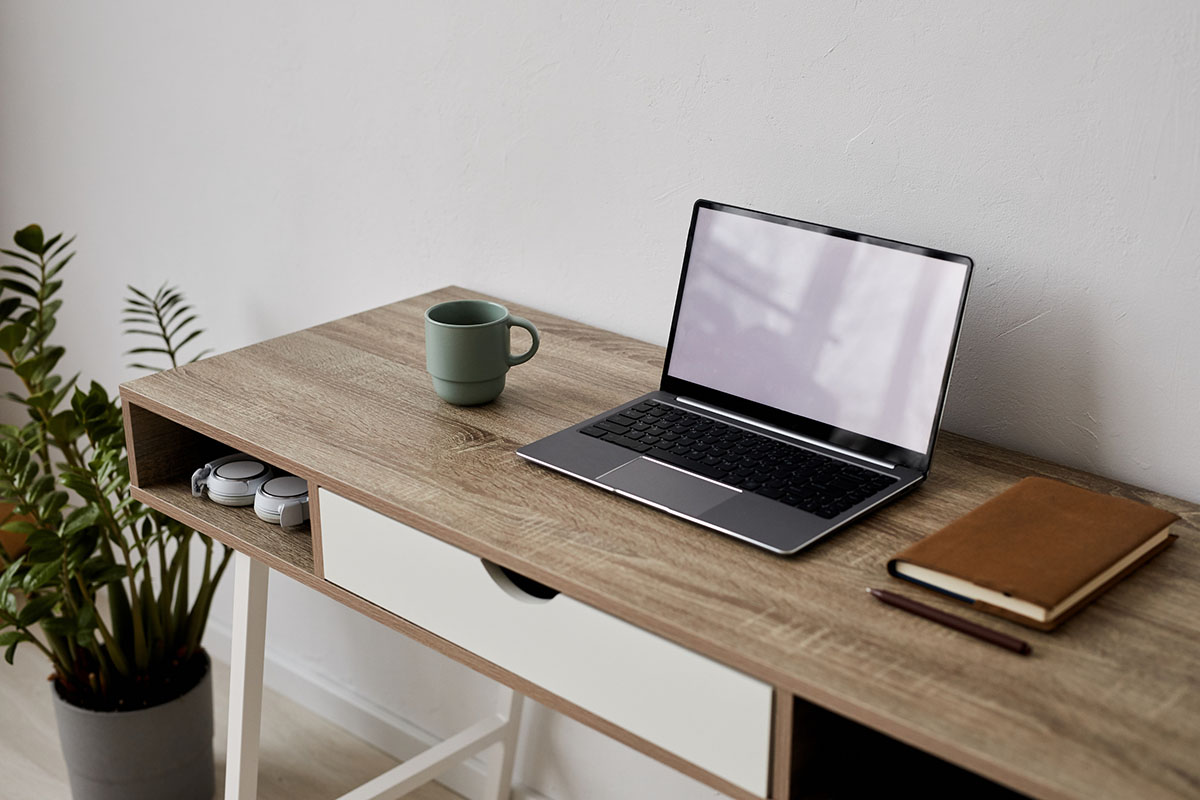 An empty home office desk with an open laptop, a coffee mug and a leather notebook