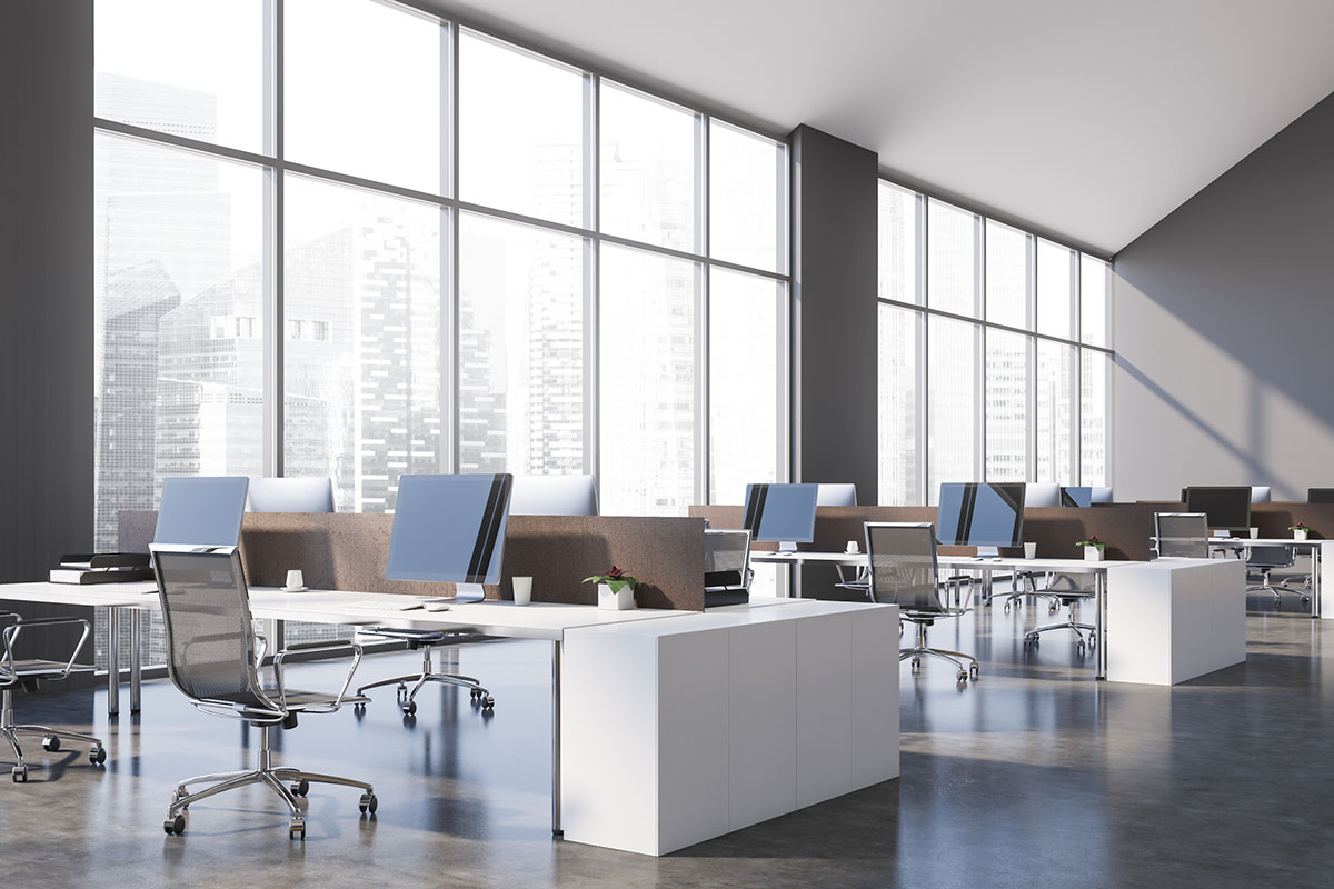 Empty open-plan office with rows of desks and monitors in front of floor-to-ceiling windows overlooking city towers