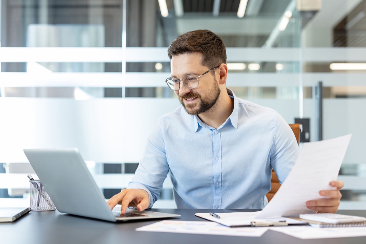 Professional in glasses reviewing a printed document while typing on a laptop at an office desk covered with paperwork
