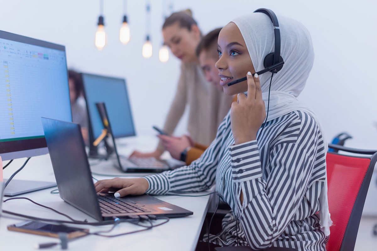 Customer service agent wearing a headset and talking with a caller while typing on a laptop in a bright office